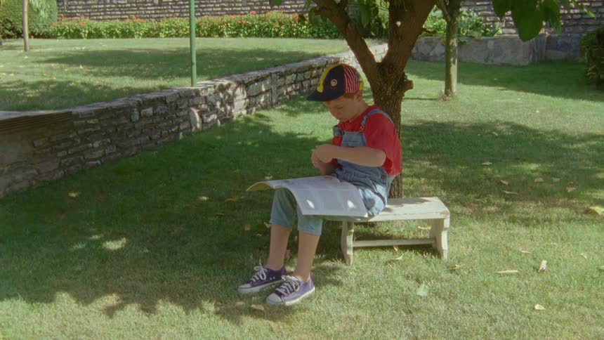 A boy is leafing through a book while sitting on a bench in the garden