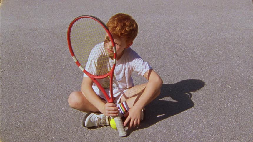 Boy sits crosslegged on asphalt looking through tennis racket
