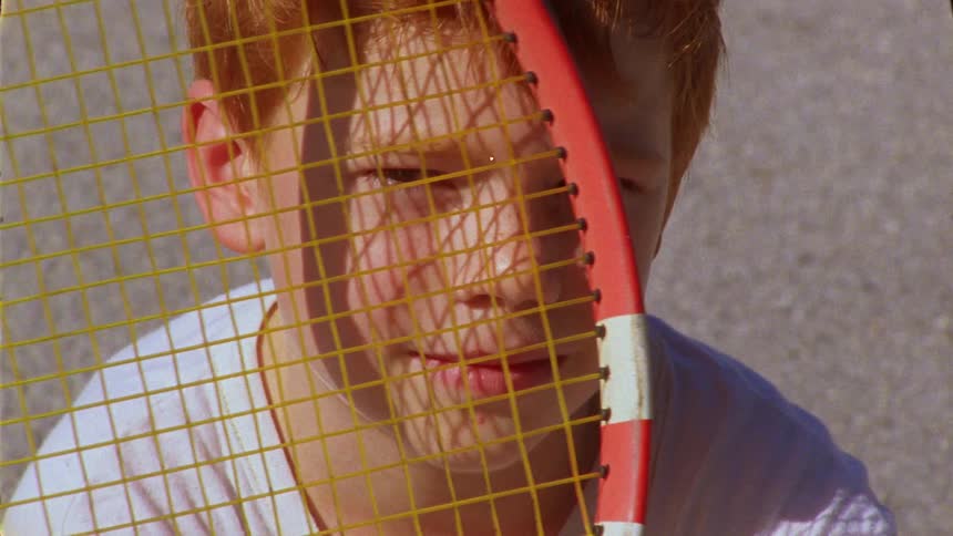 Boy holds a tennis racket up to his face while sitting on the ground during the day
