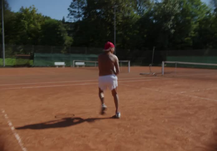 Elderly man playing tennis with another man on a court outdoors next to trees and grass during day time