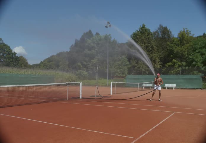 On a sunny day an elderly man uses a water hose to wet the surface of a tennis court