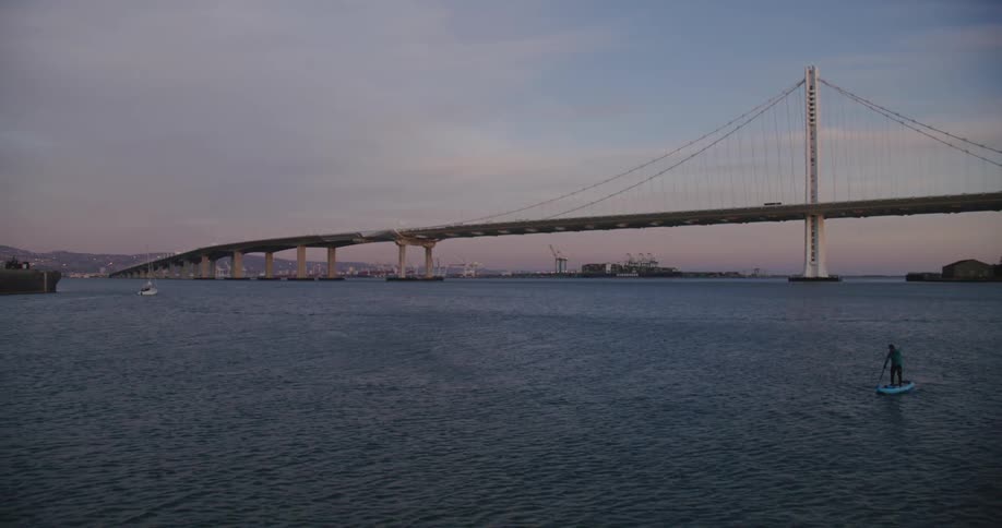 A large bridge goes across a vast lake with a person canoeing at dusk