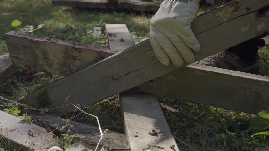 A persons gloved hand holds a wooden plank while they hammer a nail into it outside