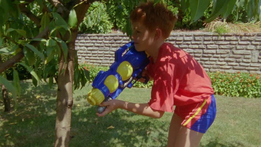A boy pumps a water gun as he hides behind a tree while his dad holds a hose