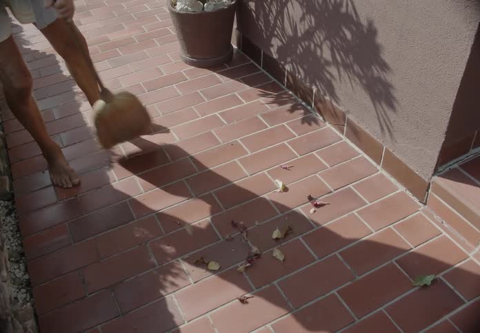 A man sweeps leaves with a broom on a brick walkway outside next to a house