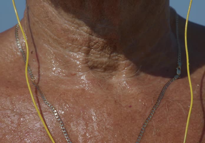 Close up of an older mans bare chest and neck wearing a necklace and yellow cords to his ears