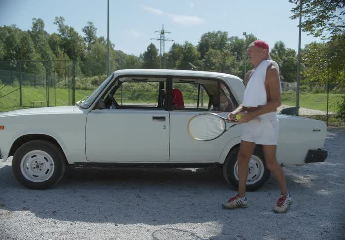 An elderly person picking up a tennis racket and tennis balls from their vehicle then closing the door and walking away