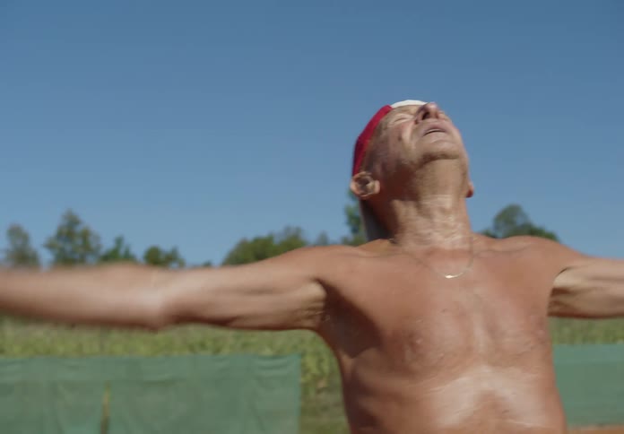A shirtless elderly man wearing a baseball cap stretches on a track with a backdrop of green vegetation