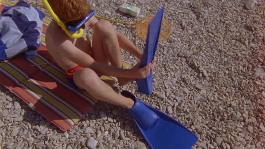 A boy puts flippers on while sitting on a stonecovered ground