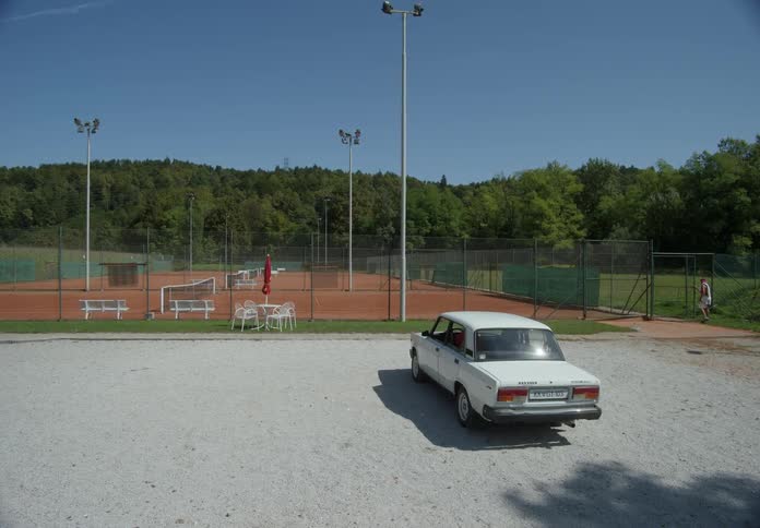 A person walking from a white sedan car into a tennis court