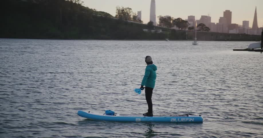Man with cap and light blue jacket on a paddle board paddling and watching city skyline at sunset