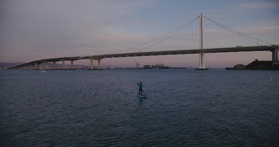 A man stands on a paddleboard paddling on the San Francisco Bay close to the Bay Bridge