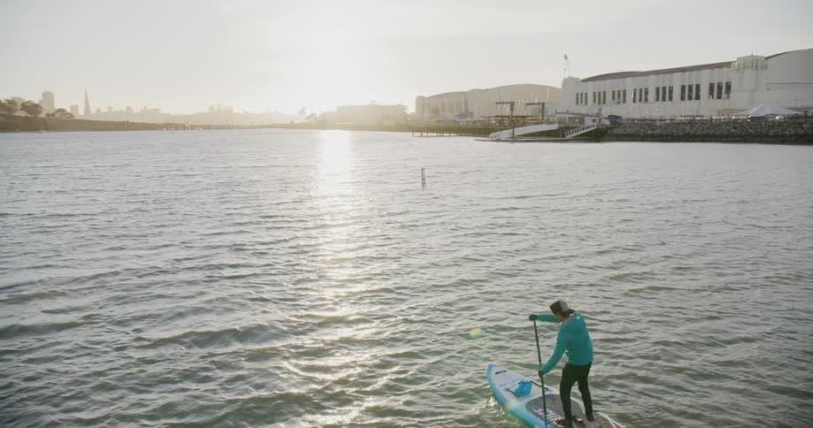 A man stand up paddle boarding on the San Francisco Bay wearing a coat in the late afternoon near sunset