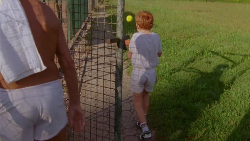 Father hands a tennis ball to his son and son runs to tennis court with racket also in hand