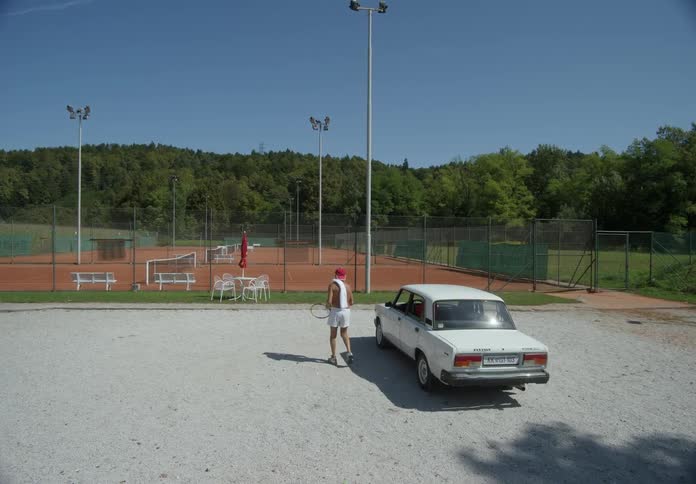 A shirtless man wearing shorts holding a tennis racket steps out of a white sedan car and heads towards a tennis court nestled amidst trees