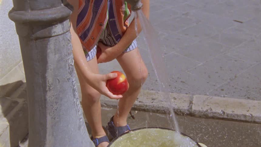 A kid washing two red apples in a tap with a bucket under