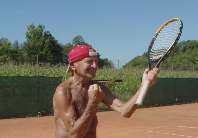 An old man with a tennis racket on a court making crooked gestures with his arm