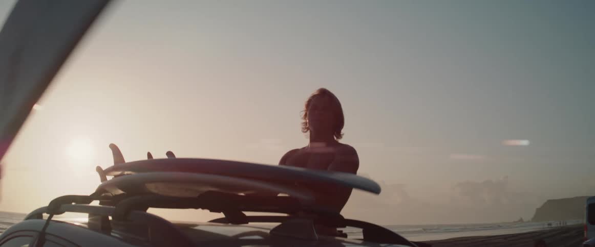 One man removes a surfboard from atop a car and hands it to his companion