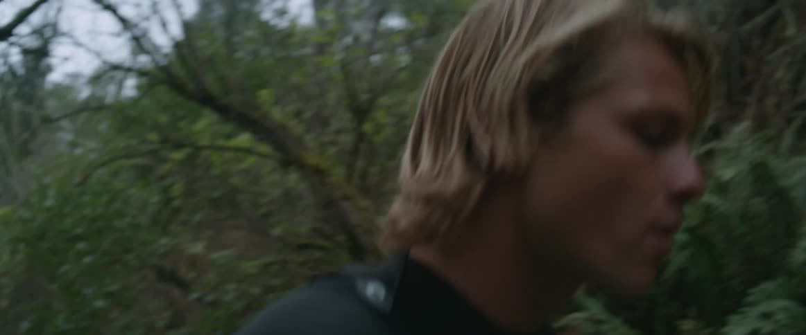 Young blonde man carries a surfboard through dense vegetation