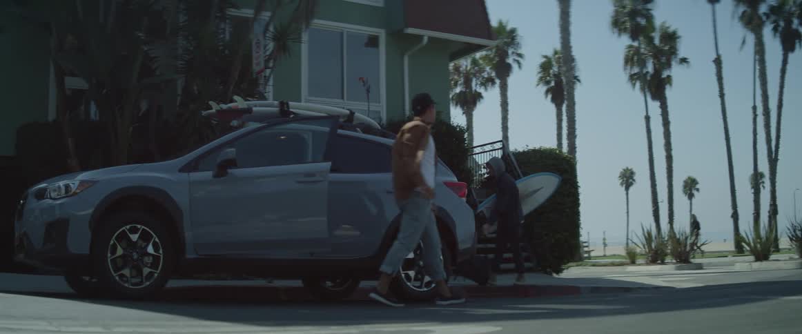 A person stepping out of an suv with another holding a surf board in front of a building surrounded by palm trees