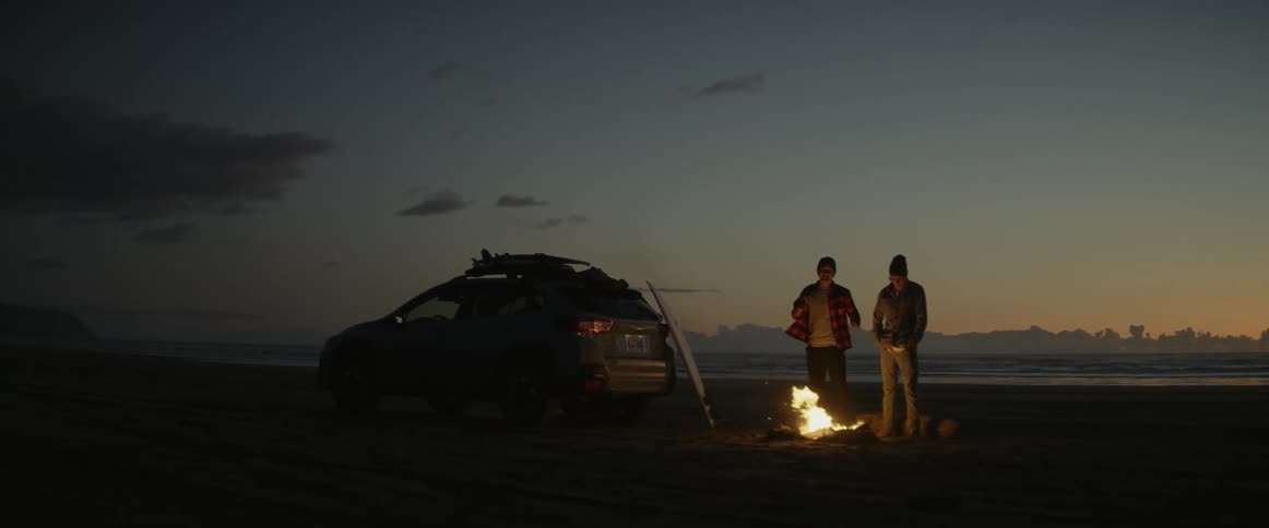 Two men facing a bonfire on the beach sand and beside them a vehicle and a surfboard