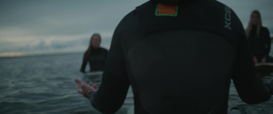People sitting on top of a surfboard in the open sea with one of them with arms open in a prayerful gesture