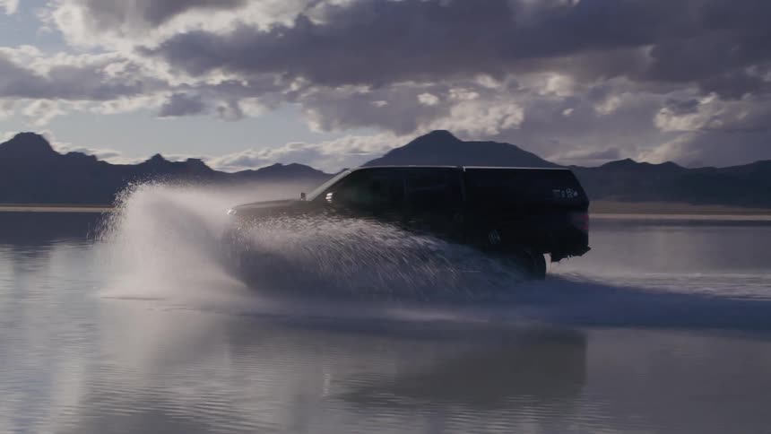 A truck driving through shallow water during sunset with a backdrop of mountains in the distance on the Salt Flats, cinema drone footage