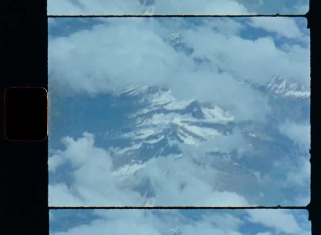 Aerial Super 8 mm footage looking out of an airplane or passenger aircraft onto an alpine mountain range such as the Alps covered in snow at high altitude and clouds