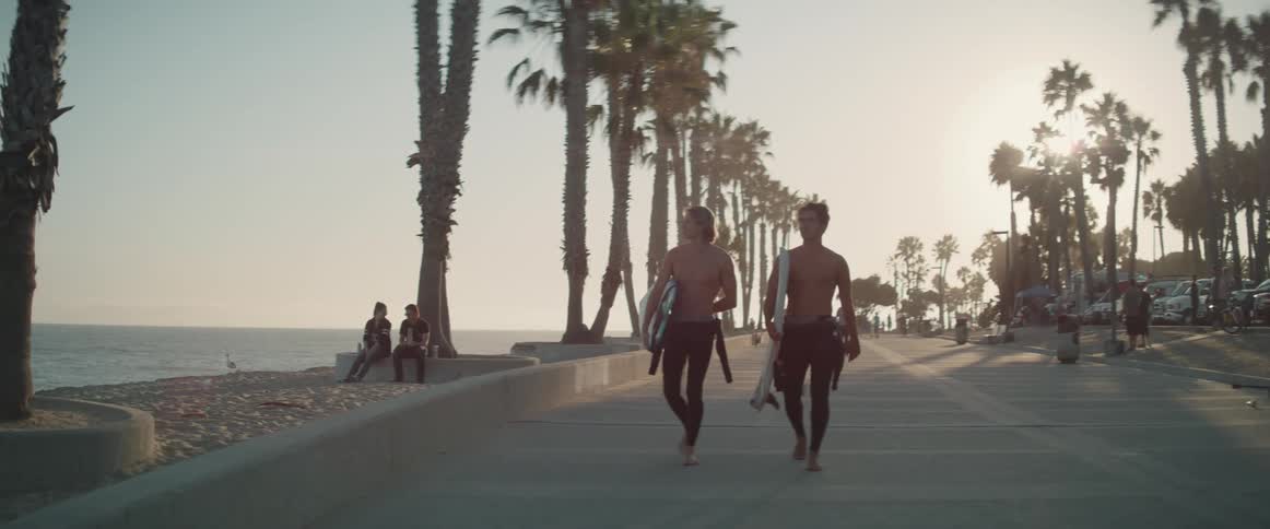 People walking and biking on a boardwalk while two men carry surfboards while talking