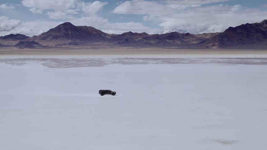 Truck races across the flooded salt flats with a background of mountains on a cloudy day, aerial footage