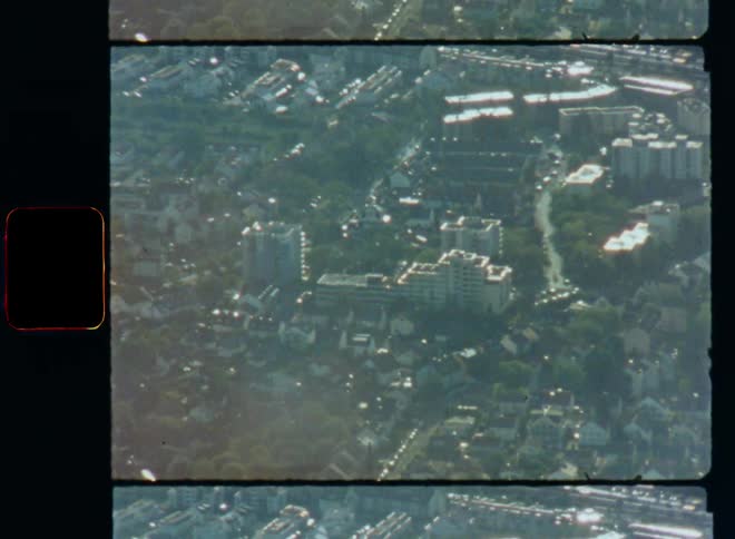 Super 8mm aerial film clip flying over a downtown city in an urban area with lots of houses large multi-story or high-rise buildings and cars driving on roads with lots of traffic in a highly populated area with developed infrastructure