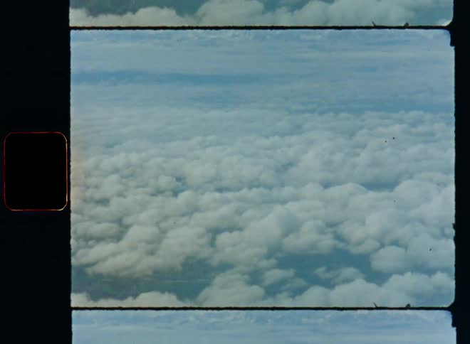 Old Super 8 film footage looking out of aircraft onto a layer of fluffy clouds hovering over the landscape on a sunny day with mountains at the horizon while flying above the clouds