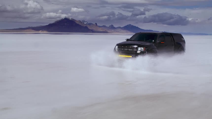 A truck drives across a flooded landscape with mountains in the background on the Salt Flats, cinema drone footage