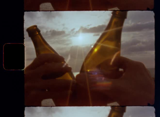 First person perspective shot of two people holding up beer bottles clinking them together as toast and celebration of summer holidays vacation while standing near the ocean on a warm and sunny day with bright sunshine and lens flare