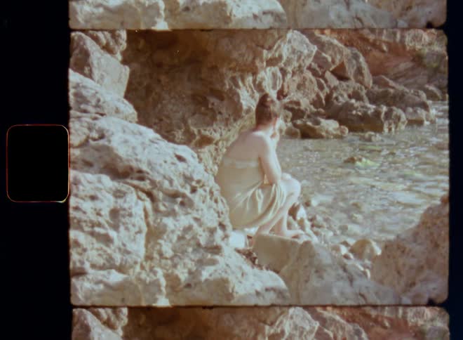 A lightly dressed sad or contemplative young woman in swimming wear sits on the rocks at the edge of the sea in a hidden secret bay or lagoon while looking at water as the waves wash up on the beach on Super 8mm film with shaky camera movement
