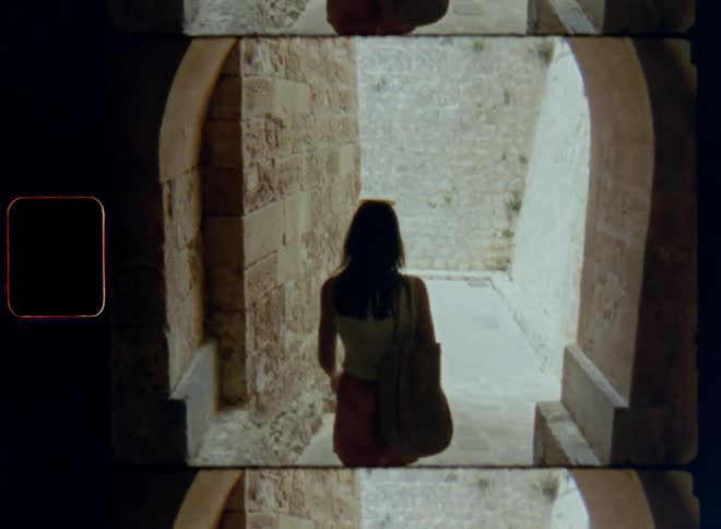 Dynamic handheld 8mm film of a woman in shorts and white top with a carrying bag walking down an alley with ancient brick or stone walls through an archway or arched gateway in an old historic town leaving shadow entering bright sun light