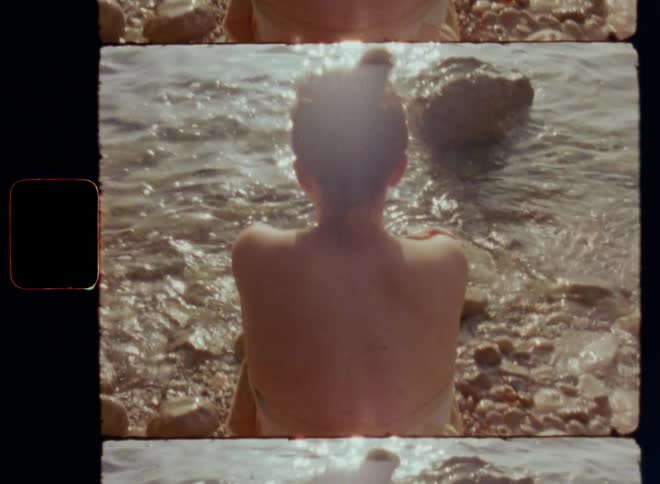 A lightly dressed young woman wrapped up in a towel sits on the beach by the water of the sea at dawn looking into the moving water with bright light reflections reminiscing and thinking on her own or alone in nature feeling the elements on S 8mm