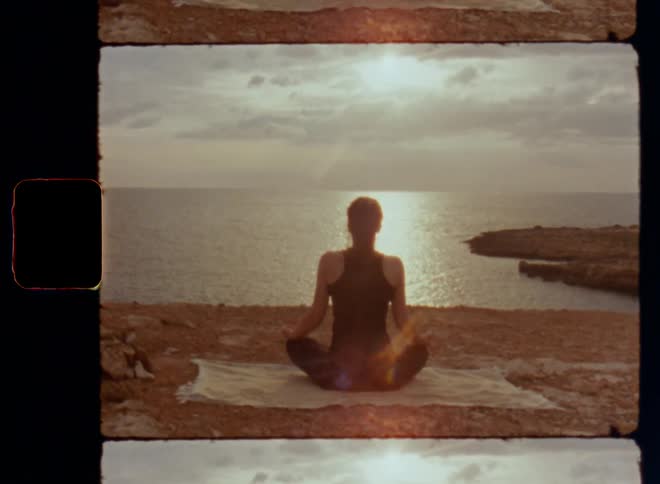Tilting Super 8mm shot of a spiritual young woman in black clothing sitting cross-legged doing yoga or meditation on rocks facing the ocean and horizon as the bright sun is warming her body and heart with positive vibes energy and inner peace on 8mm film