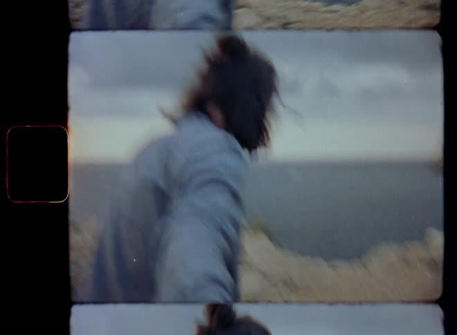A young man with long brown hair smiles and is happy while taking and holding the hand of another person while walking towards the edge of the cliff next to the sea filmed on Super 8mm film in a first person view perspective