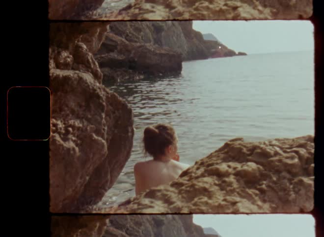 Pretty naked woman with brown hair and bun hairstyle sits at base of rocks and cliffs near water looking out to sea enjoying a peaceful and quite day or evening in a hidden bay by the beach as the waves ripple and move on Super 8mm film