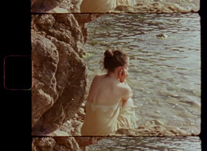 Brunette woman wearing yellow strapless dress or towel sits on narrow beach near coastal rocks and cliffs with her head rested on her arms while looking at moving water with a rippling surface and sunlight reflections on her own or alone on Super 8mm
