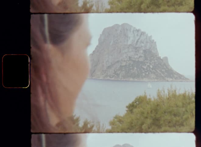 Young woman looks at island next to the coast and enjoying view from the high cliff on vintage Super-8 film revealing a spectacular rock mountain in the water and a sailing boat on the ocean exploring the coastline or shoreline of the Mediterranean Sea