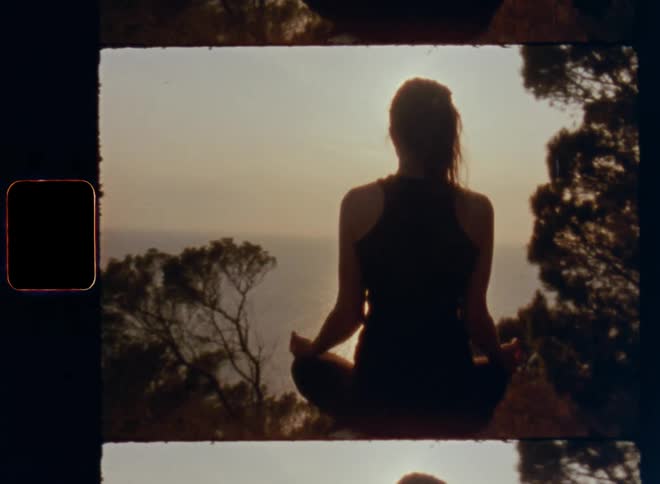 Panning Super 8 shot with lens flare due to bright sunshine during sunset of alternative open-minded spiritual girl meditating while sitting cross-legged on a mountain overlooking the sea or horizon connecting to herself and nature in peaceful atmosphere