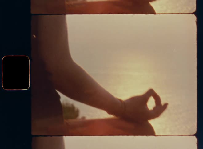 Vintage 8 mm close up of a woman sitting crosse-legged with her hands on her knees meditating at the beach or coast overlooking the sunset over the ocean or sea with camera movement and bright glaring sunlight and lens flare