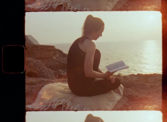 Handheld dynamic 8mm film footage walking towards a woman sitting on a beige blanket reading a book on a cliff edge overlooking the ocean as the sun sets in the sea while she relaxes and enjoys a great story and the peaceful and harmonic atmosphere