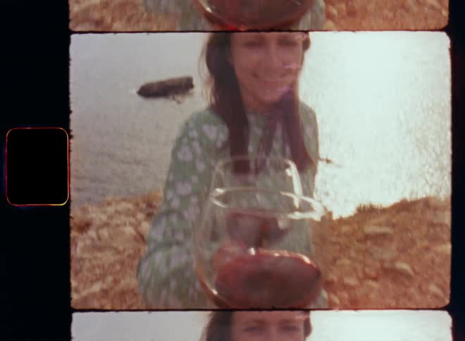 Friends or love couple consisting of brunette woman with long hair in a green dress and an unrecognisable person filming on vintage S8 film from point of view shot perspective toasting with glasses of red wine on the seashore at sunset