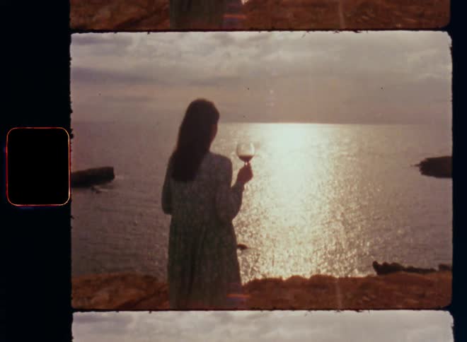 Backside of a woman holding wine glass while she overlooks the sunlight on the ocean before she turns around and looks into old vintage Super 8 camera with lens flare and glare caused by the bright light and the reflection on the water