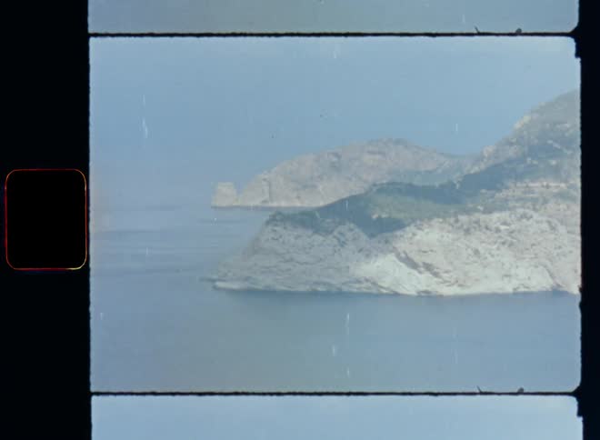 An aerial view shot on vintage Super 8mm camera of a peninsula or island with rocky stone mountains in the middle of the Mediterranean Sea or ocean surrounded by blue water and the clouds at the bright horizon