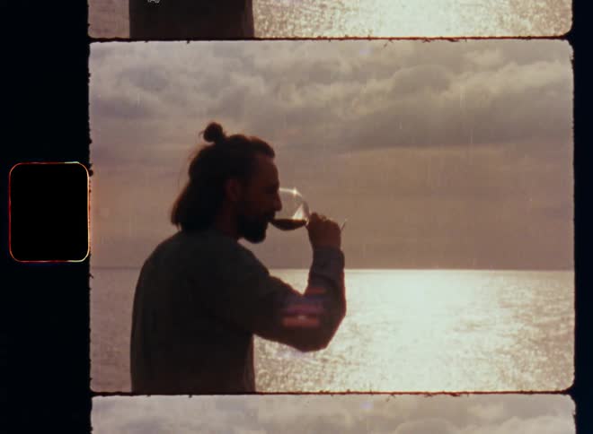Handheld shaky 8mm film footage of the dark silhouette of a man drinking red wine on the beach against the bright cloudy sky with large clouds at sunset facing the sea or a lake with a bright light reflection in the background
