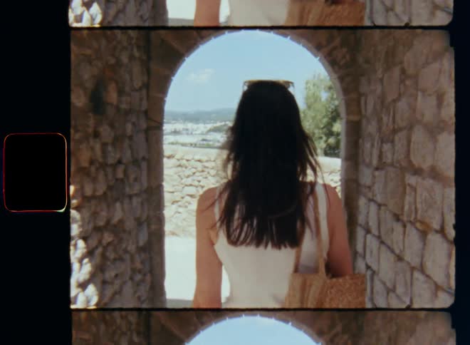 Young brunette lady with lose brown long hair carrying a handbag walking out of a ruined building or archway made of cobble stone to exit or enter a place through the entrance or doorway of an old house or castle in daytime on a sunny day
